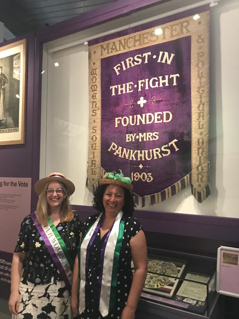 Maya and Helen in front of suffragette banner saying "First in the  fight | Founded by Mrs Pankhurst | 1903
